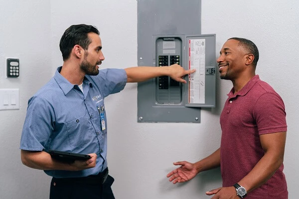 A Mr. Electric service professional holding a tablet pointing at an open electrical panel while talking to a smiling man