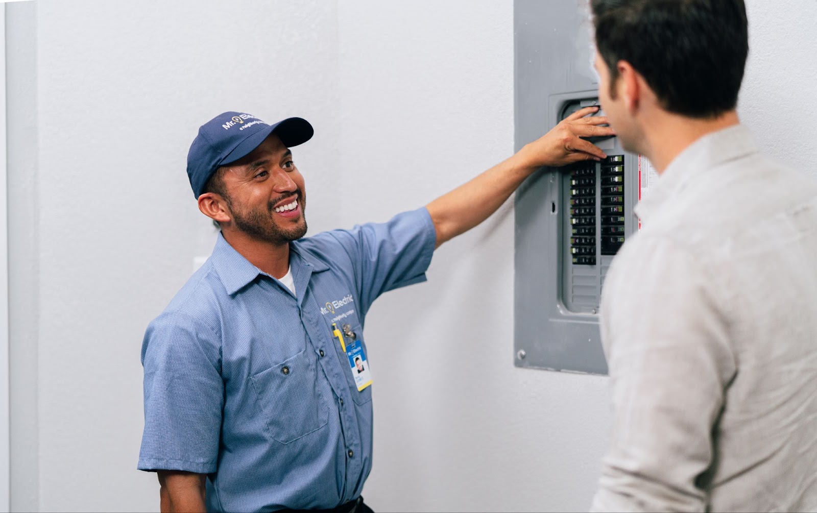 A mre techniciansmiles while pointing at an electrical panel, as he explains something to a person.