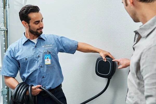 An electrician holds a cord attached to an EV charger on a wall and shows another man how to use it.