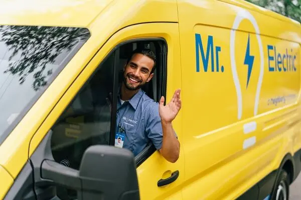  A smiling Mr. Electric electrician in a yellow Mr. Electric van waves his arm out of the window.