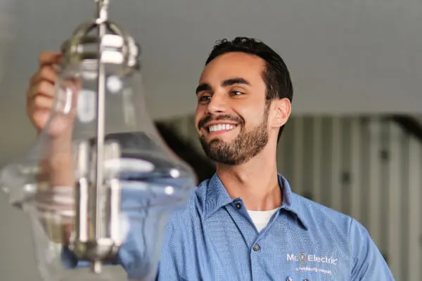  A smiling Mr. Electric electrician reaches out to a pendant light fixture suspended from a ceiling.