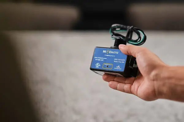 A hand holds a black house surge protector with coiled cords on the top and a blue label with a Mr. Electric logo on it.