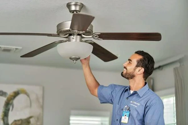 A smiling Mr. Electric electrician reaches over a light fixture attached to a ceiling fan