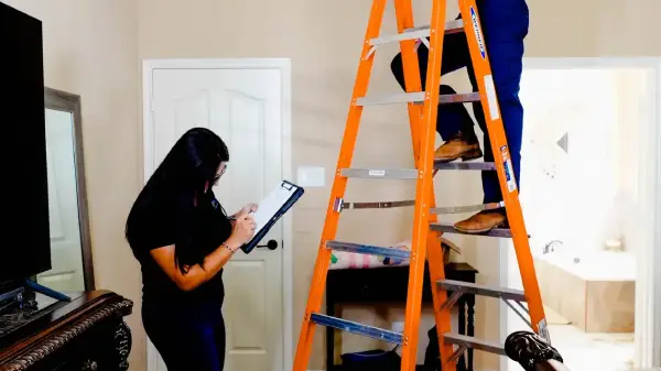 male electrician on a ladder installing a smoke detector while female electrician writes on clipboard