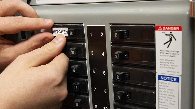 A person adds a white label that says Kitchen to a black circuit breaker on a gray electrical panel.