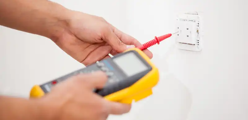 Person's right hand holding a multimeter while the left hand holds a meter probe to an electrical outlet.