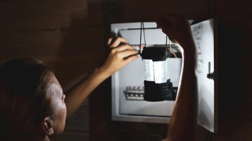 Woman holding a battery power lantern checking a circuit panel during a power outage.