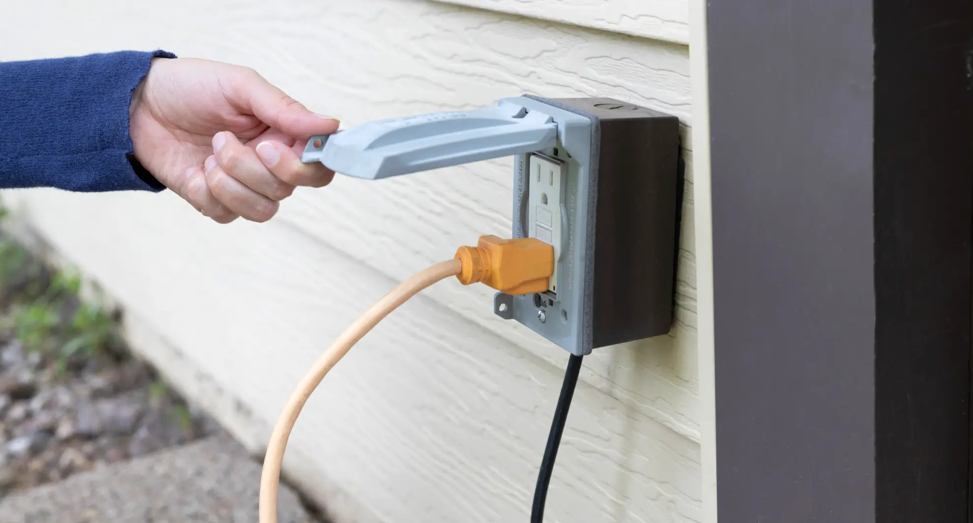 A hand lifting an outlet cover for an outdoor plug.