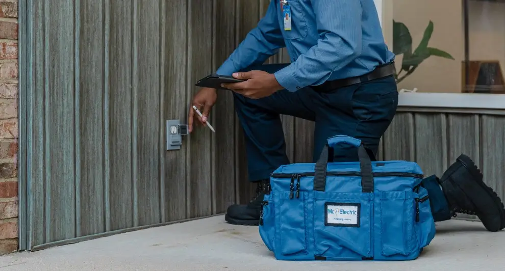 Electrician in a blue uniform kneeling outdoors to inspect a GFCI electrical outlet mounted on an exterior wall, with a blue Mr. Electric service bag in the foreground, preparing for an electrical safety inspection or repair.