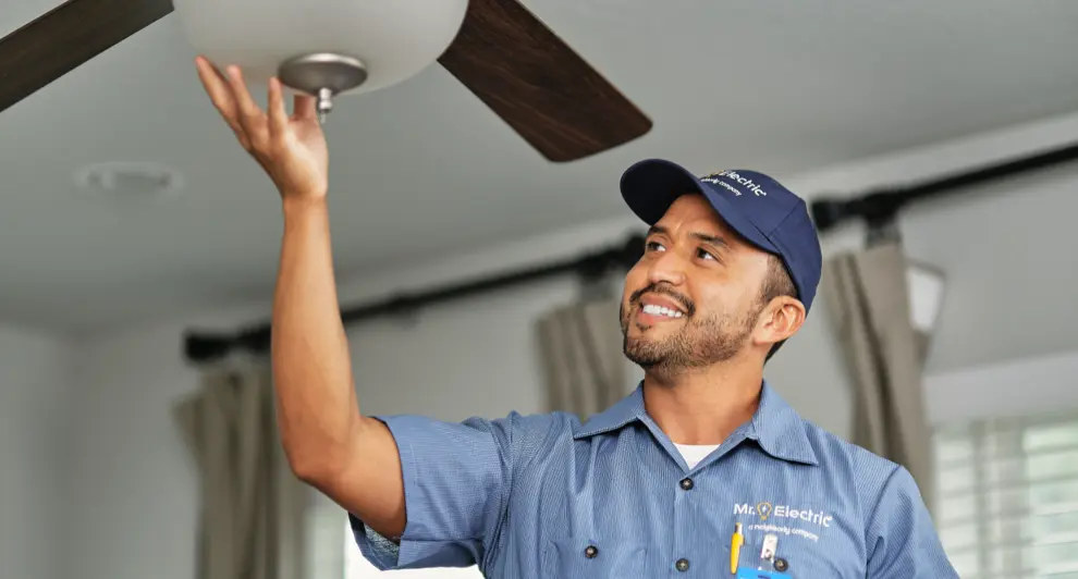 A professional electrician in a blue uniform and cap is installing a ceiling fan