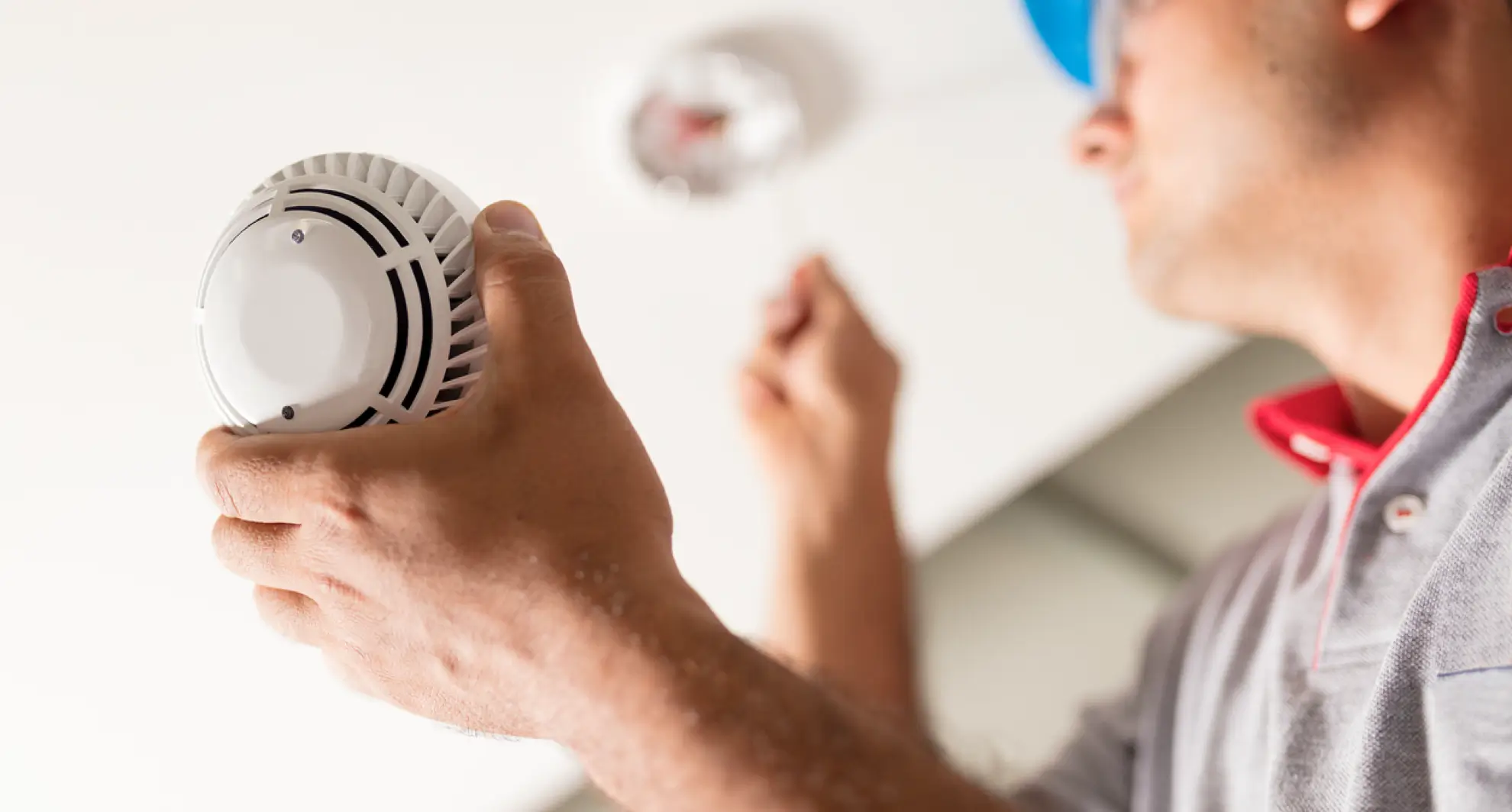 Close-up of a male technician in a light-gray work polo with red trim and a blue hard hat, holding a white smoke detector in one hand while using a screwdriver to secure the mounting bracket to a smooth white ceiling; focus is on his hands and the device.