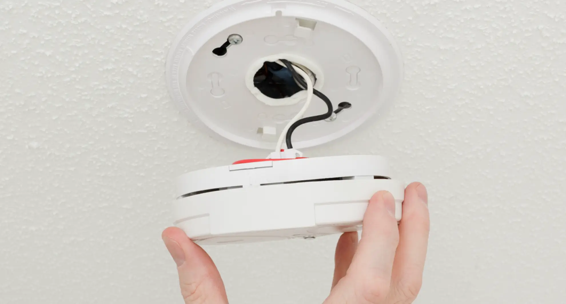 Detailed view of a hand attaching the base of a hardwired smoke alarm to a textured white ceiling; black and white wires emerge from the ceiling box as the alarm housing is lifted into place.