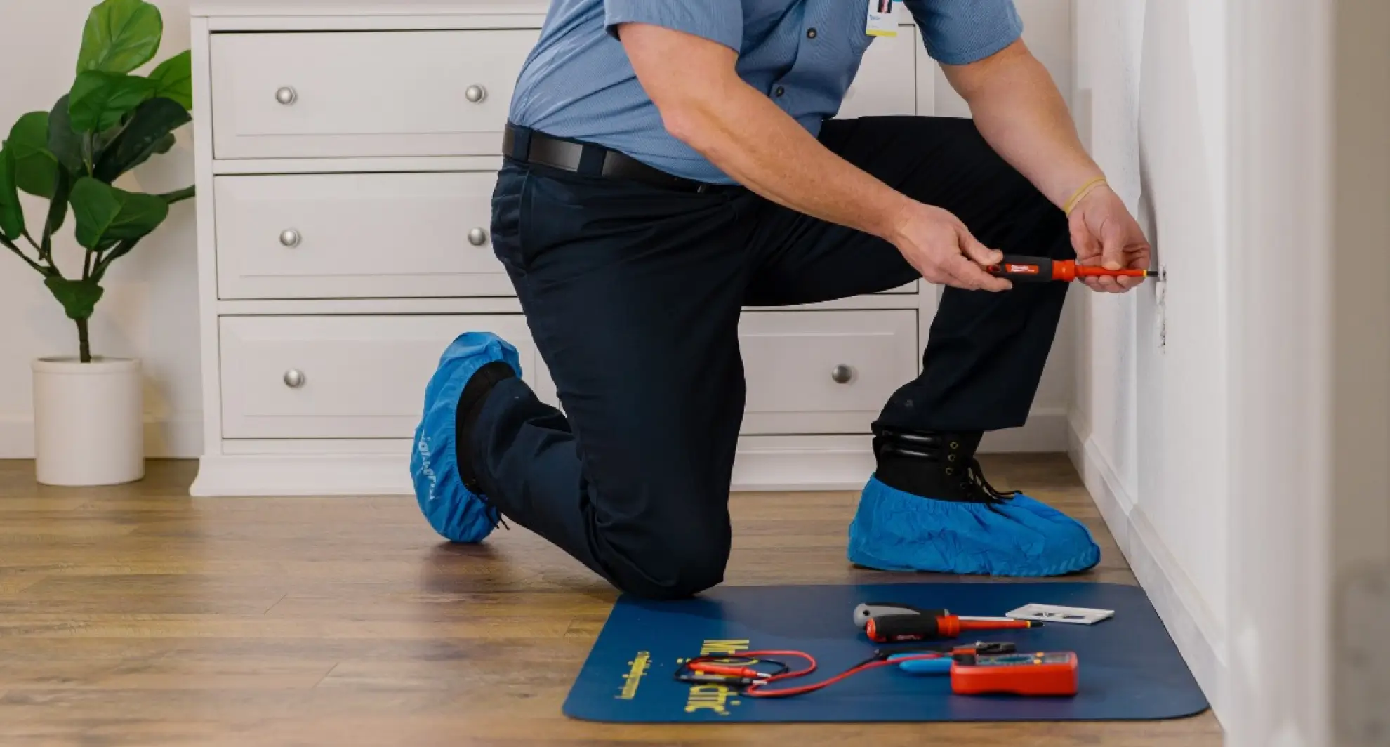 Indoor scene of a uniformed technician in a light-blue short-sleeve shirt and navy work pants kneeling on one knee, wearing black boots covered by bright-blue disposable shoe covers, using an insulated orange screwdriver to service a wall outlet; a white dresser, potted plant, and neatly arranged tools on a blue floor mat rest on a hardwood floor in the background.