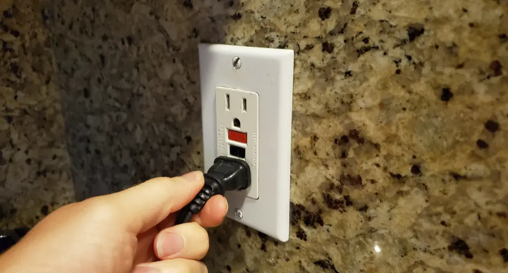 A close-up image of a hand plugging a black cord into a GFCI outlet on a speckled granite backsplash.
