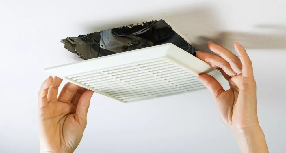 Hands are seen installing a bathroom exhaust fan cover into a ceiling. The open vent reveals wiring and the fan mechanism, emphasizing its role in ventilation.