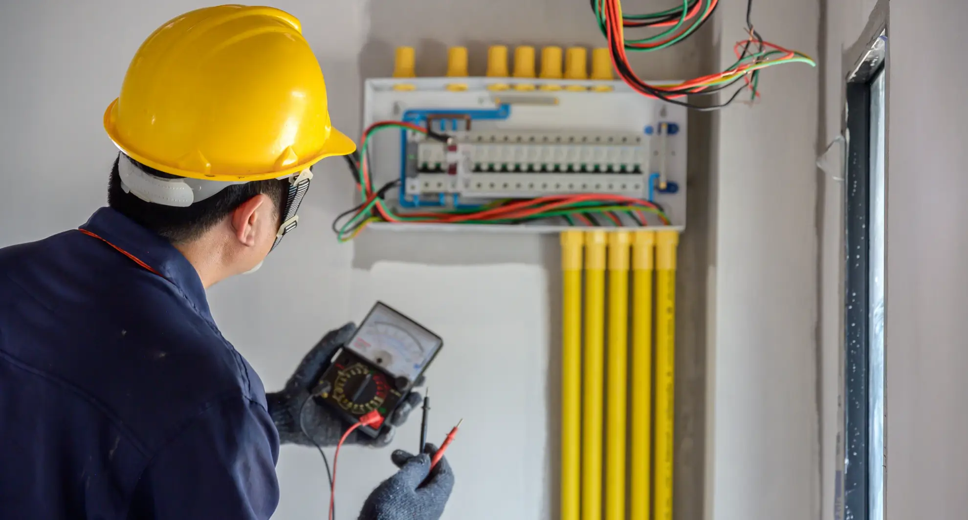 Electrician in yellow hard hat testing a home electrical panel with a multimeter