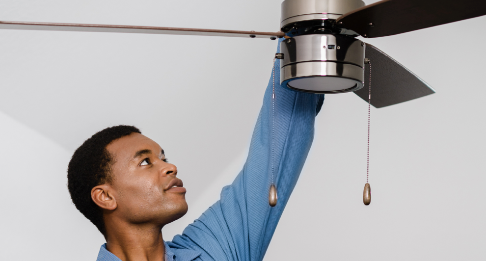 Man in blue shirt carefully installing an indoor ceiling fan with pull chains.