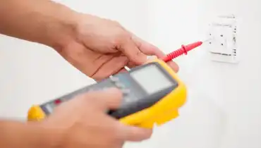 Person's right hand holding a multimeter while the left hand holds a meter probe to an electrical outlet.